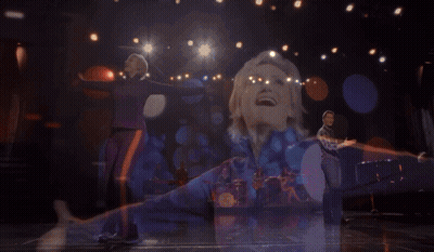 Woman In Purple And Orange Jacket Singing Into Microphone GIF