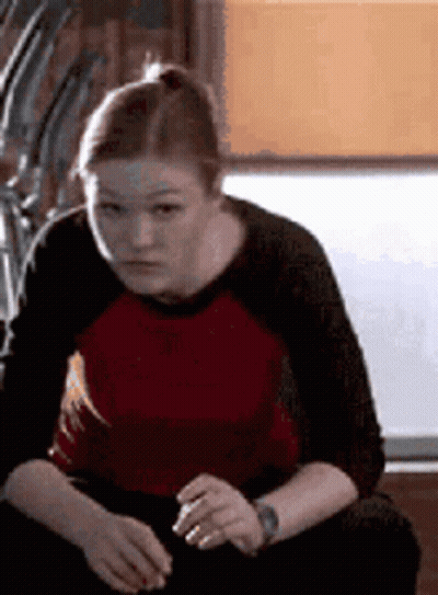 Woman In Red And Black Shirt Sitting On Couch Looking At Camera GIF