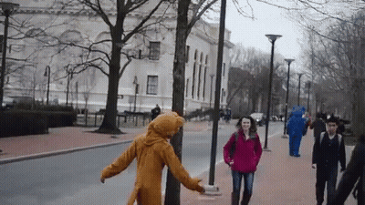 Woman In Red Jacket Walking With Man In Bear Costume Behind GIF