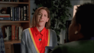 Woman In Red Shirt And Grey Sweater Waving At Another Woman In Front Of Bookshelf GIF