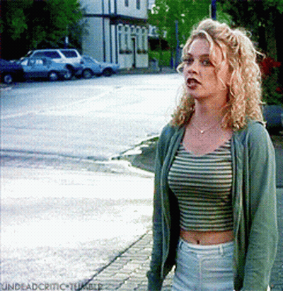 Woman In Striped Crop Top And Green Jacket On Sidewalk GIF
