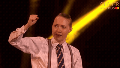 Woman In Striped Shirt And Suspenders Holding Fist Up Before Olivier Awards Light GIF
