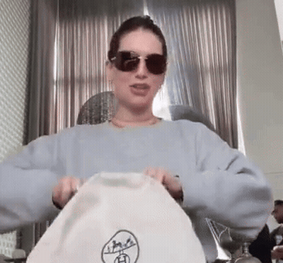 Woman In Sunglasses And Blue Sweater Holding White Bag GIF