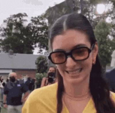 Woman In Sunglasses And Yellow Shirt Smiling Before Crowd GIF