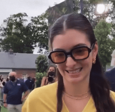 Woman In Sunglasses And Yellow Shirt Smiling Before Crowd GIF