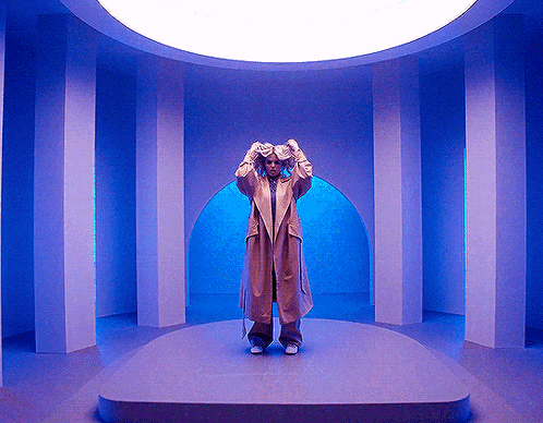 Woman In Trench Coat Standing In Room With Blue Columns GIF