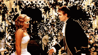 Woman In White Dress And Mask Next To Man In Black Suit GIF