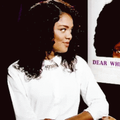 Woman In White Shirt Sitting In Front Of Dear White Sign GIF