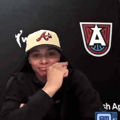 Woman In Yellow Hat And Black Hoodie Sitting In Front Of Wall With Logo GIF