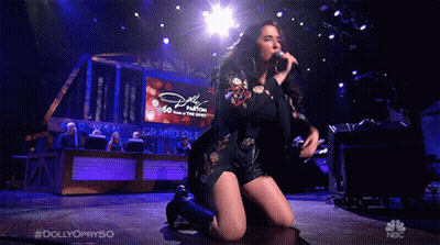 Woman Kneeling Singing Into Microphone In Front Of Dolly Parton Screen GIF