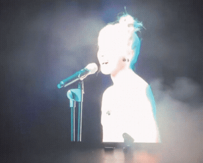 Woman Singing Into Mic On Stage With Screen Behind GIF