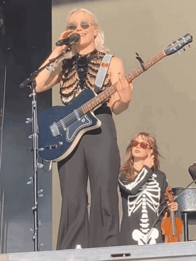 Woman Singing Into Microphone Holding Guitar Beside Girl In Skeleton Outfit GIF