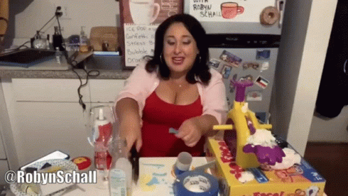 Woman Sitting At Table In Front Of Fridge With Robyn Schall Sign GIF