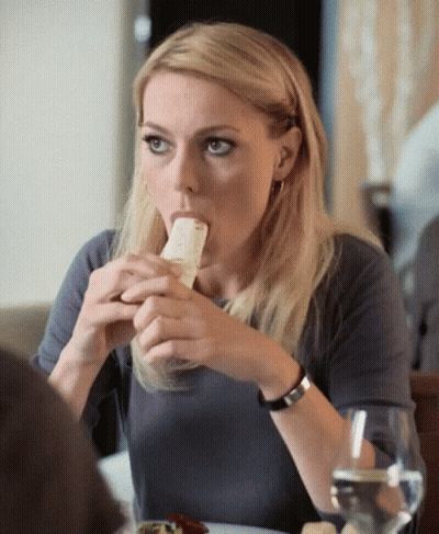 Woman Sitting At Table With Sandwich In Mouth GIF