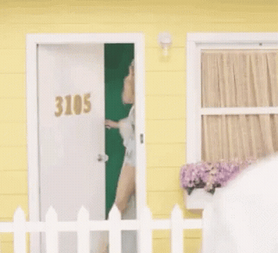 Woman Standing In Front Of Yellow House Behind White Picket Fence GIF