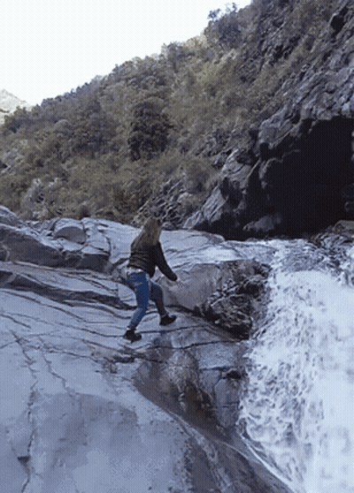 Woman Walking Down Rocky Cliff Near Waterfall GIF