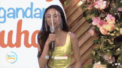 Woman With Face Shield Speaking Into Microphone In Front Of Ganda Sign GIF