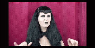 Woman With Long Black Hair And Black Makeup Making Funny Face In Front Of Red Curtain GIF