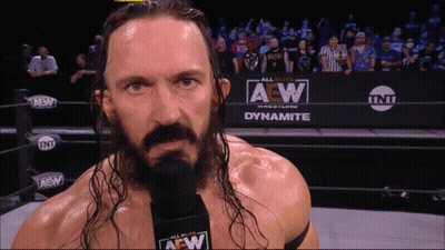 Wrestler With Long Hair And Microphone In Front Of Dynamite Sign GIF