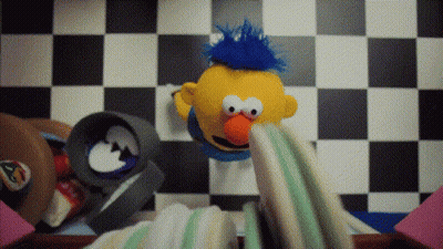 Yellow Puppet With Blue Mohawk Sitting On Checkered Floor GIF
