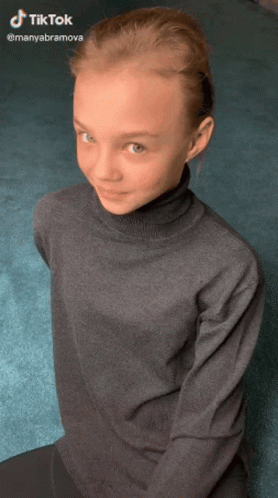 Young Girl In Turtleneck Sitting On Floor Looking At Camera GIF