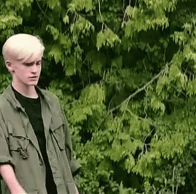 Young Man In Green Jacket And Black Shirt Standing Before Tree GIF