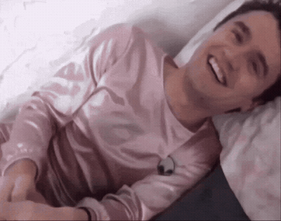 Young Man In Pink Shirt Laying On Bed Smiling GIF