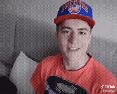 Young Man In Red Shirt And Blue Hat Sitting On Couch GIF