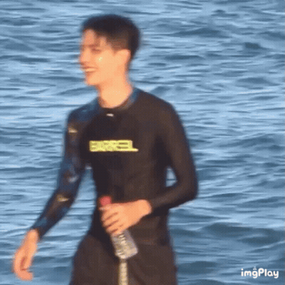 Young Man In Wetsuit Walking On Beach Holding Water GIF