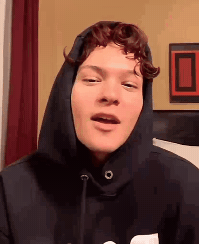 Young Man With Curly Hair In Black Hoodie Talking To Camera GIF