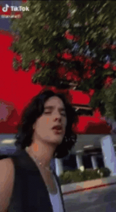 Young Man With Long Hair And Pearl Necklace In Front Of Red Flower GIF