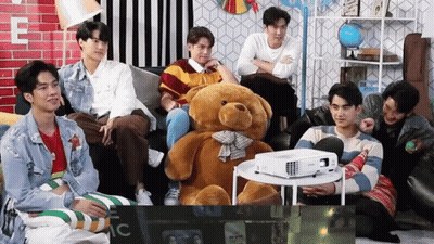 Young Men Sitting Around Teddy Bear And Projector GIF