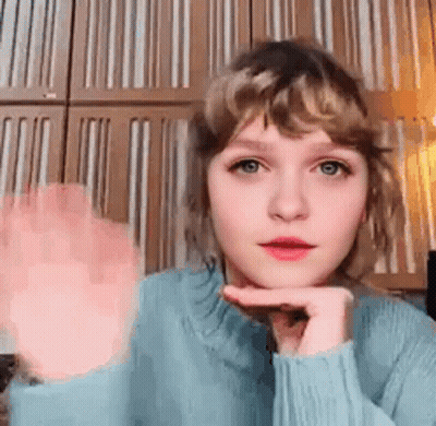 Young Woman In Blue Sweater Waving Hand Near Striped Wall GIF
