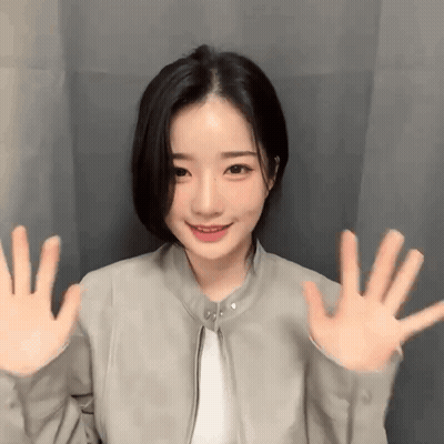 Young Woman In Gray Jacket Waving Hands By Curtain GIF