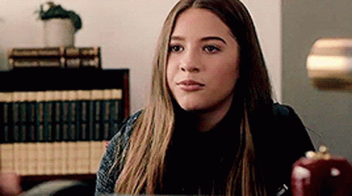 Young Woman Sitting At Desk In Front Of Bookshelf Looking At Camera GIF
