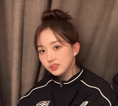 Young Woman With Hair Bun Sitting In Front Of Curtain GIF